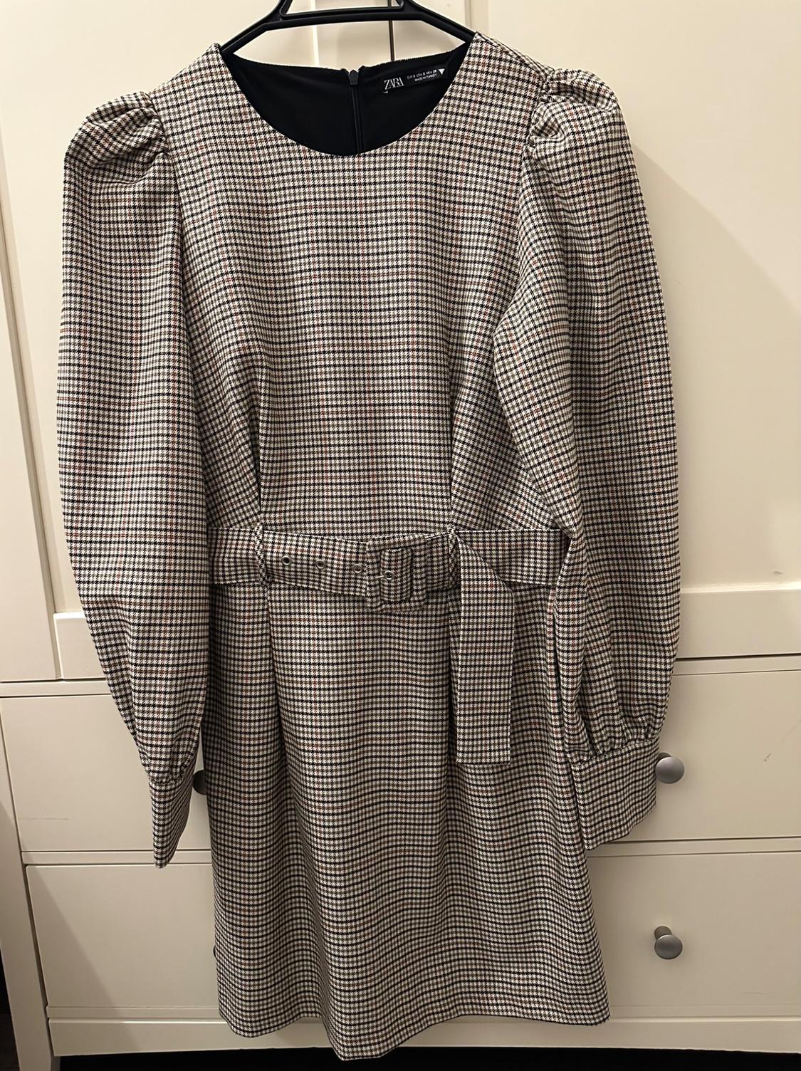 Small checked dress in brown and black tones, with puff sleeves and a belt, hanging on a hanger in front of a white wardrobe.
