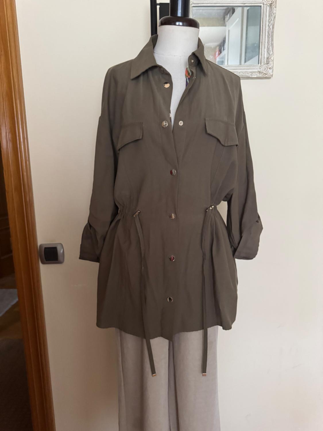 Mannequin wearing an olive green button-up jacket with adjustable drawstrings, over beige trousers. Interior background with mirror and door.