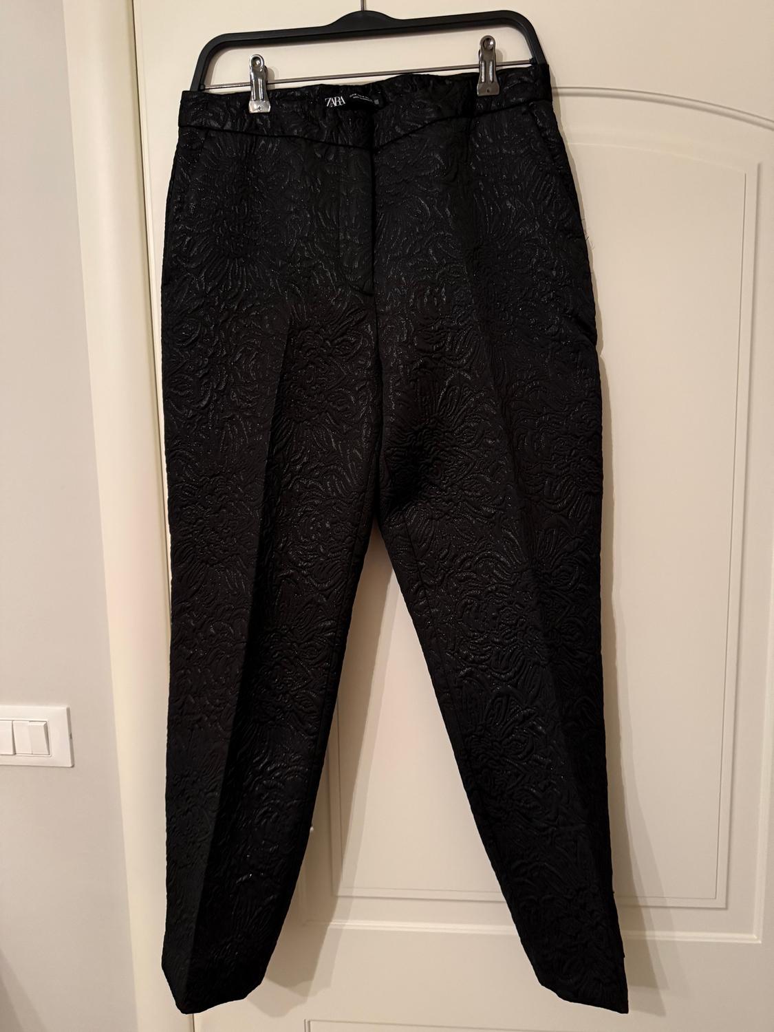 Black trousers with a floral texture, hanging on a black hanger over a white door. Label visible on the waistband.