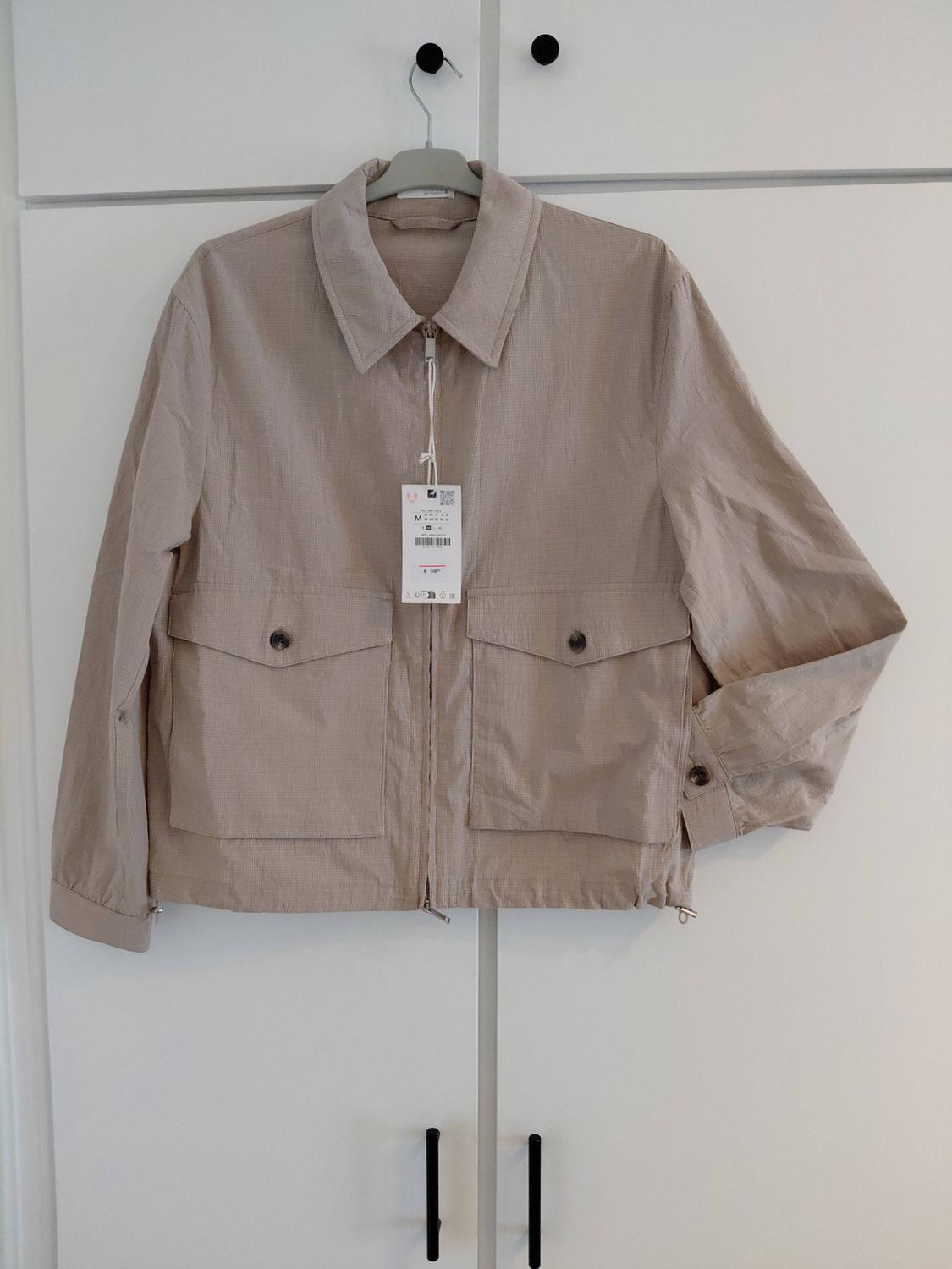 Beige long-sleeved jacket with a zip and front pockets, hanging on a coat hanger over the doors of a white wardrobe.
