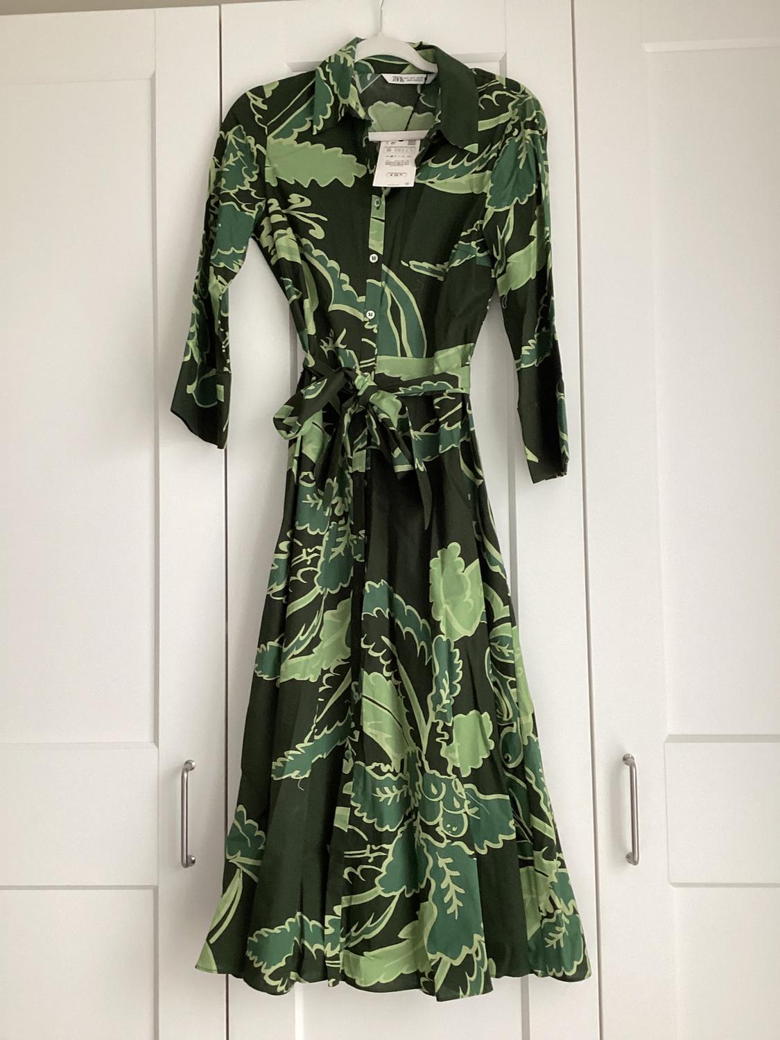 Green dress with a leaf print in light and dark shades, hanging on a white door. Label visible on the collar.
