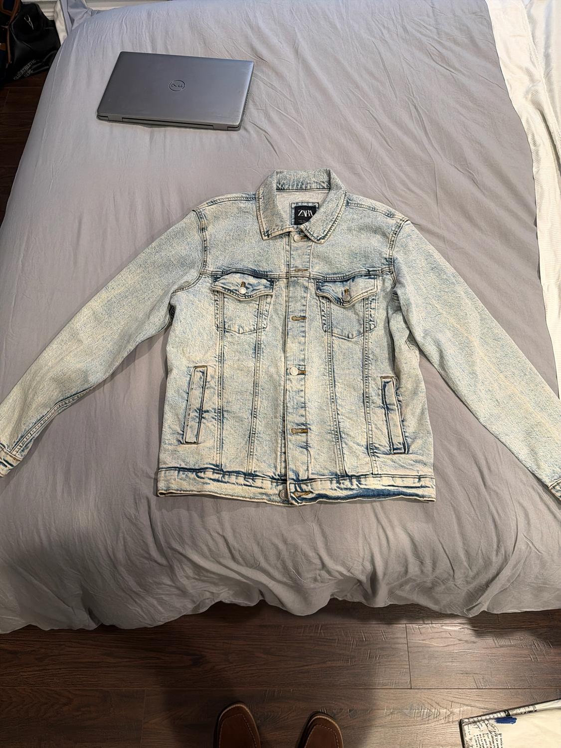 Light blue distressed denim jacket on a grey bed. Relaxed setting with a laptop and shoes peeking into view. Text on label: ZARA.