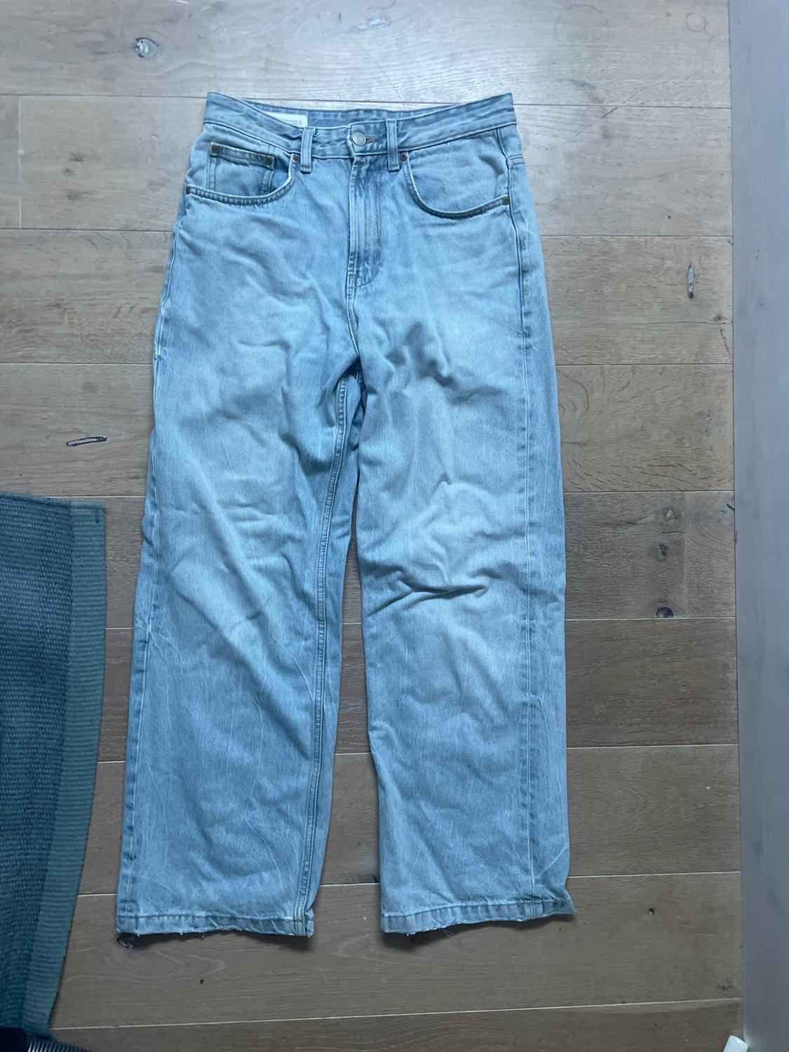 Light-blue wide-leg denim trousers laid out on a wooden floor, with the edge of a green rug on the left.