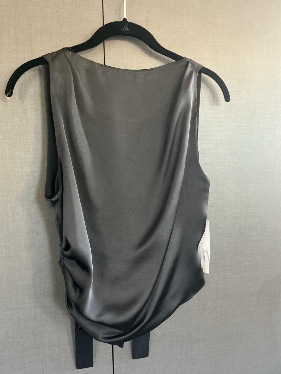 Black satin sleeveless top with draped neckline and side ruching detail, hanging on a black hanger against a textured gray background.