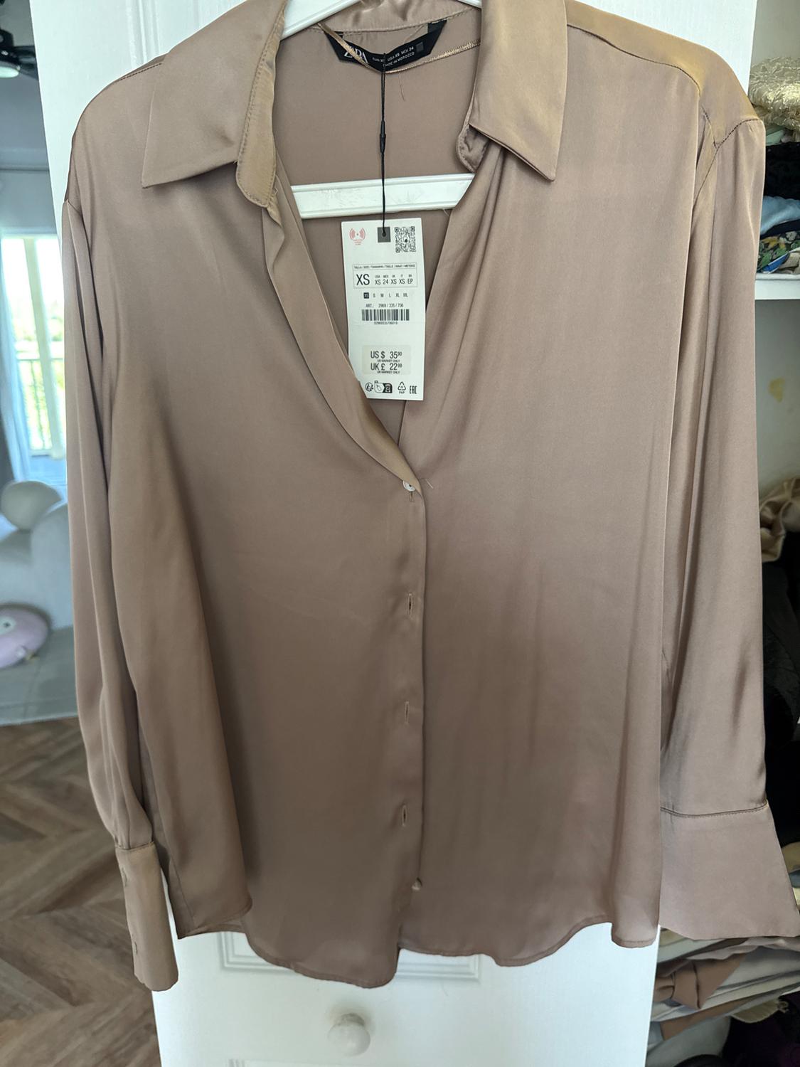 Beige satin shirt hanging on a white hanger, with price tag visible. Room background illuminated by window.