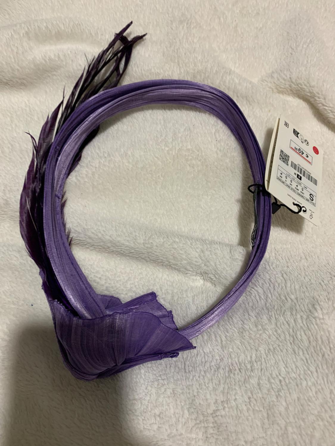 Purple headband with feathers on white fabric. Visible tag with barcode and price. Elegant and striking style.