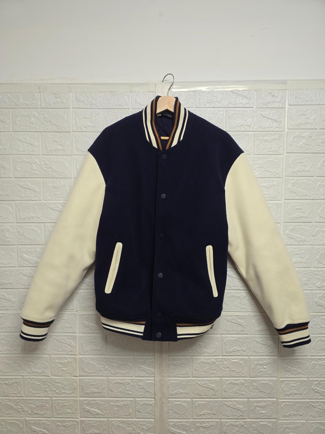 Varsity jacket hanging on a white tiled wall. Dark blue body, cream-white sleeves and detailing.