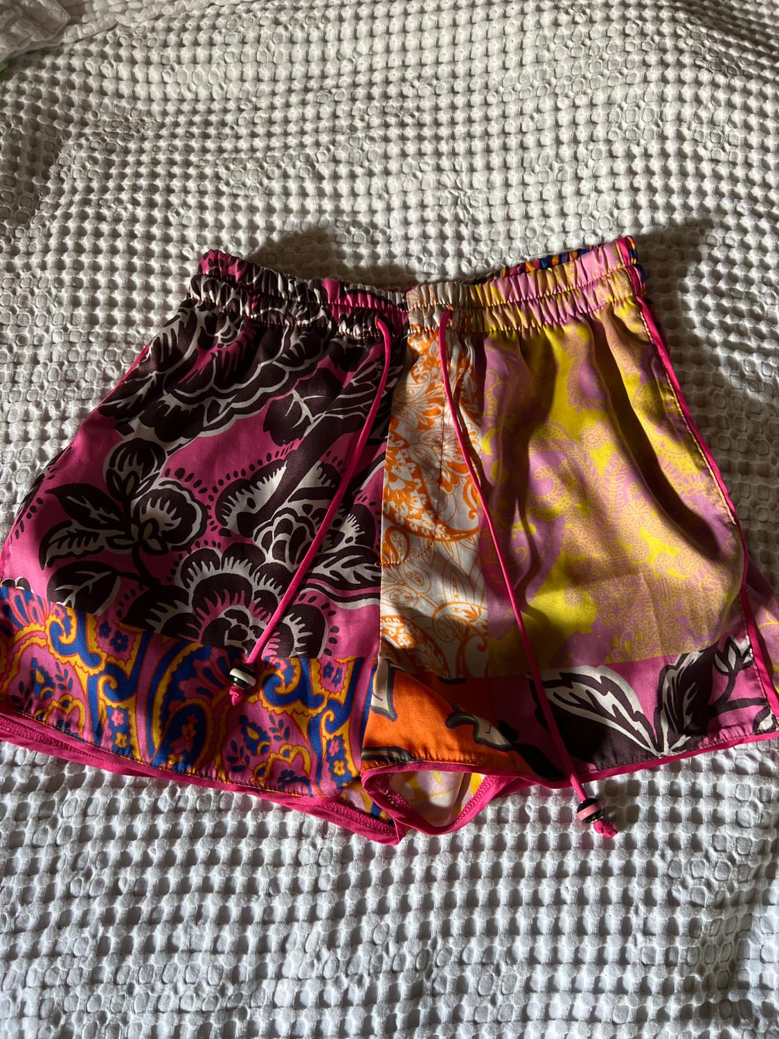 Multicoloured patchwork-print shorts with floral motifs. Elasticated waist with a drawstring, on a white textured fabric.