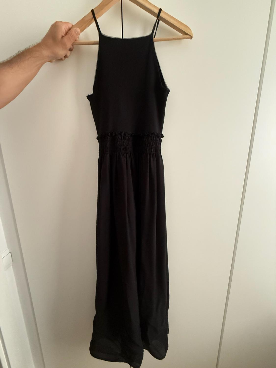 Hand holding a wooden hanger with a long black slip dress with delicate straps and a gathered elasticated waist against a white background.