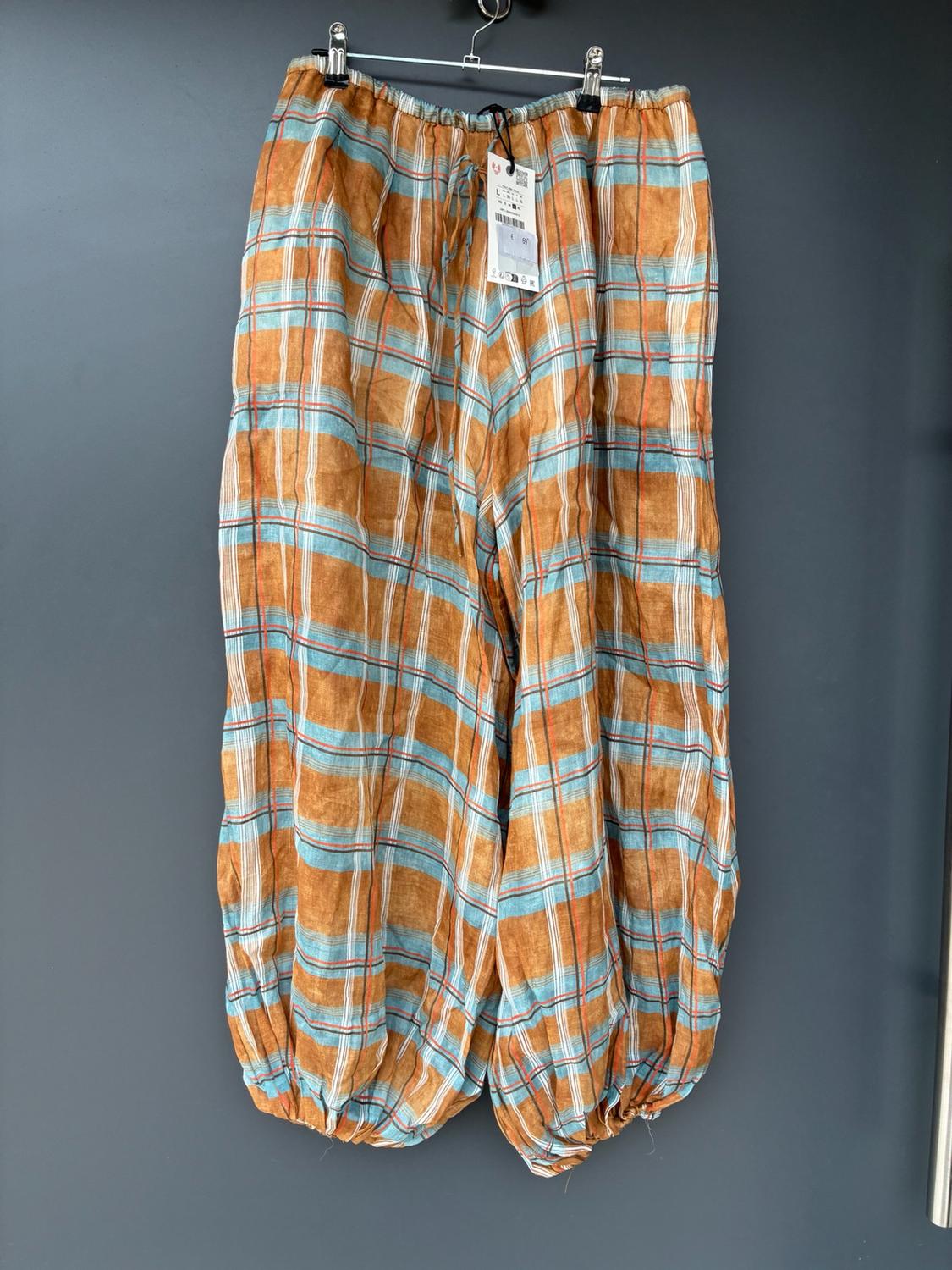 Wide-leg checked trousers in shades of orange, blue and white hanging on a hanger against a grey background. Elasticated waist and ankles.