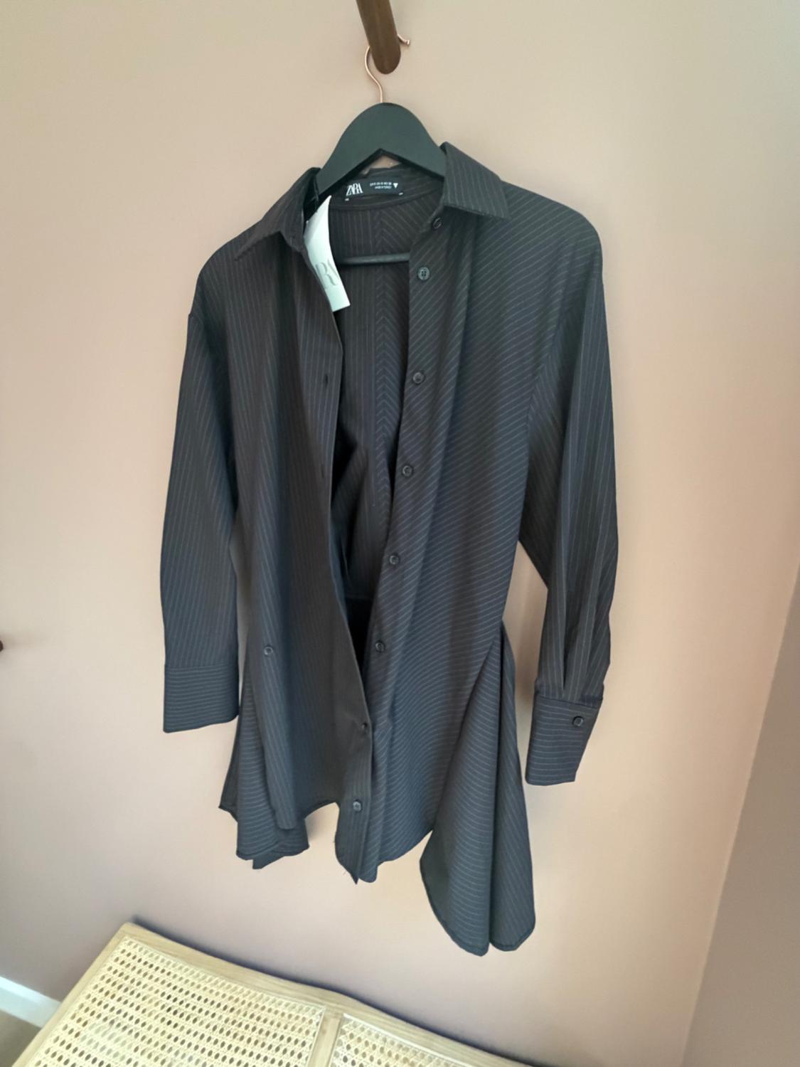 Dark striped shirt hanging on a black hanger against a beige wall. ZARA label visible on the collar. Lattice furniture underneath.
