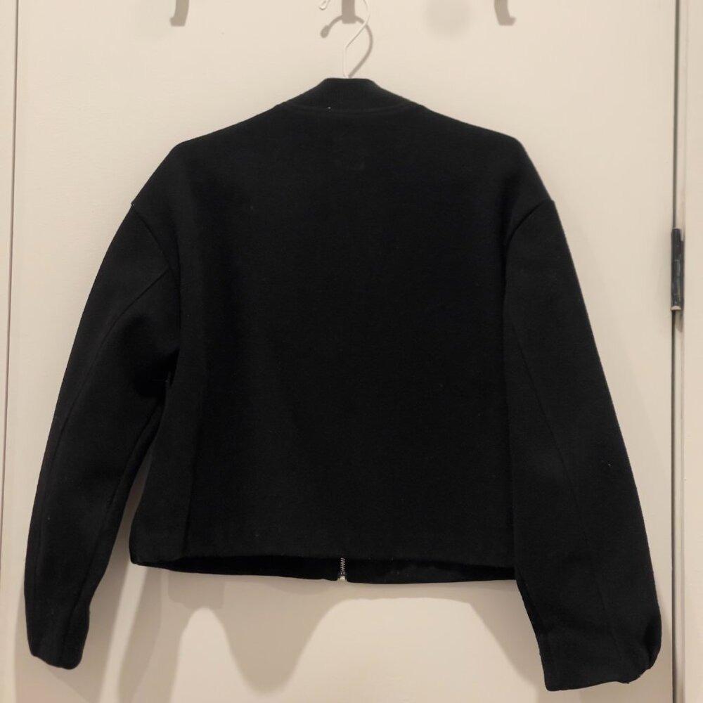 Black jacket with a cropped cut and bomber style, seen from the back, hanging on a hanger on a white door.