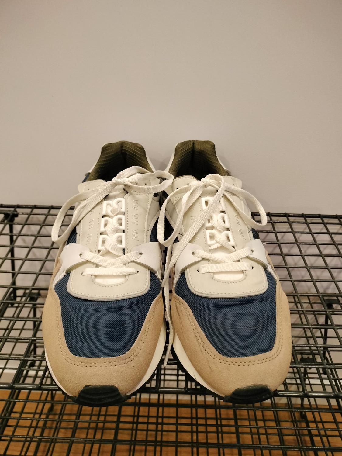 Beige, navy blue and white trainers with laces, viewed from the front on a black metal wire rack.