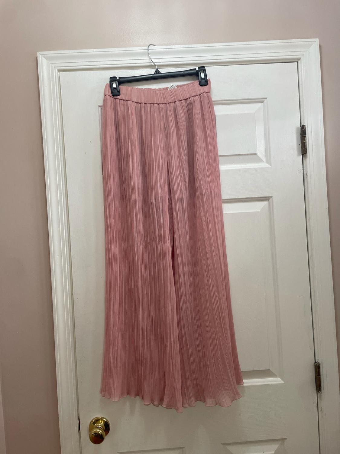 Wide-leg pink pleated pants hanging on a hanger over a white door with a gold doorknob in a home setting.
