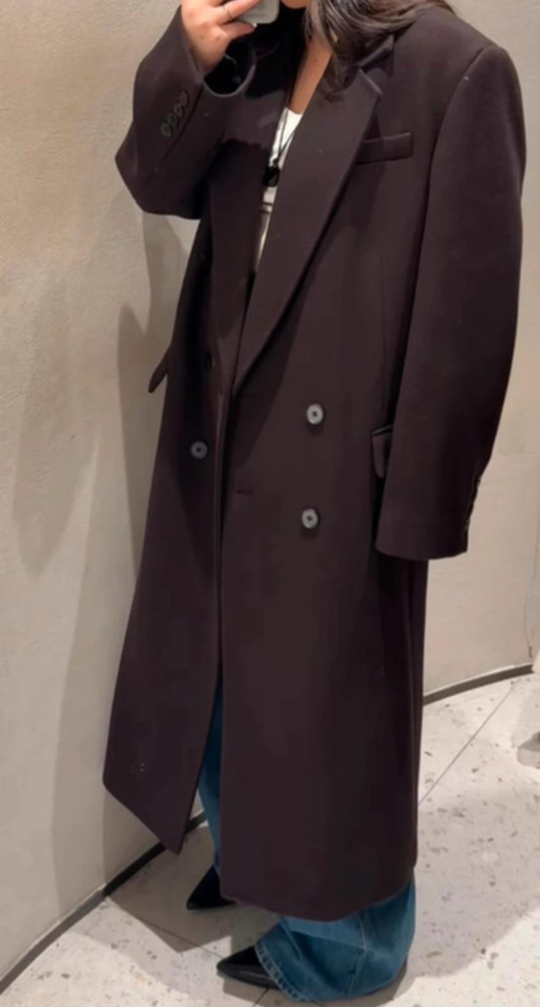 Woman in a long black coat, wide-leg jeans, and black pointed shoes, taking a photo in a mirror in a minimalist setting.