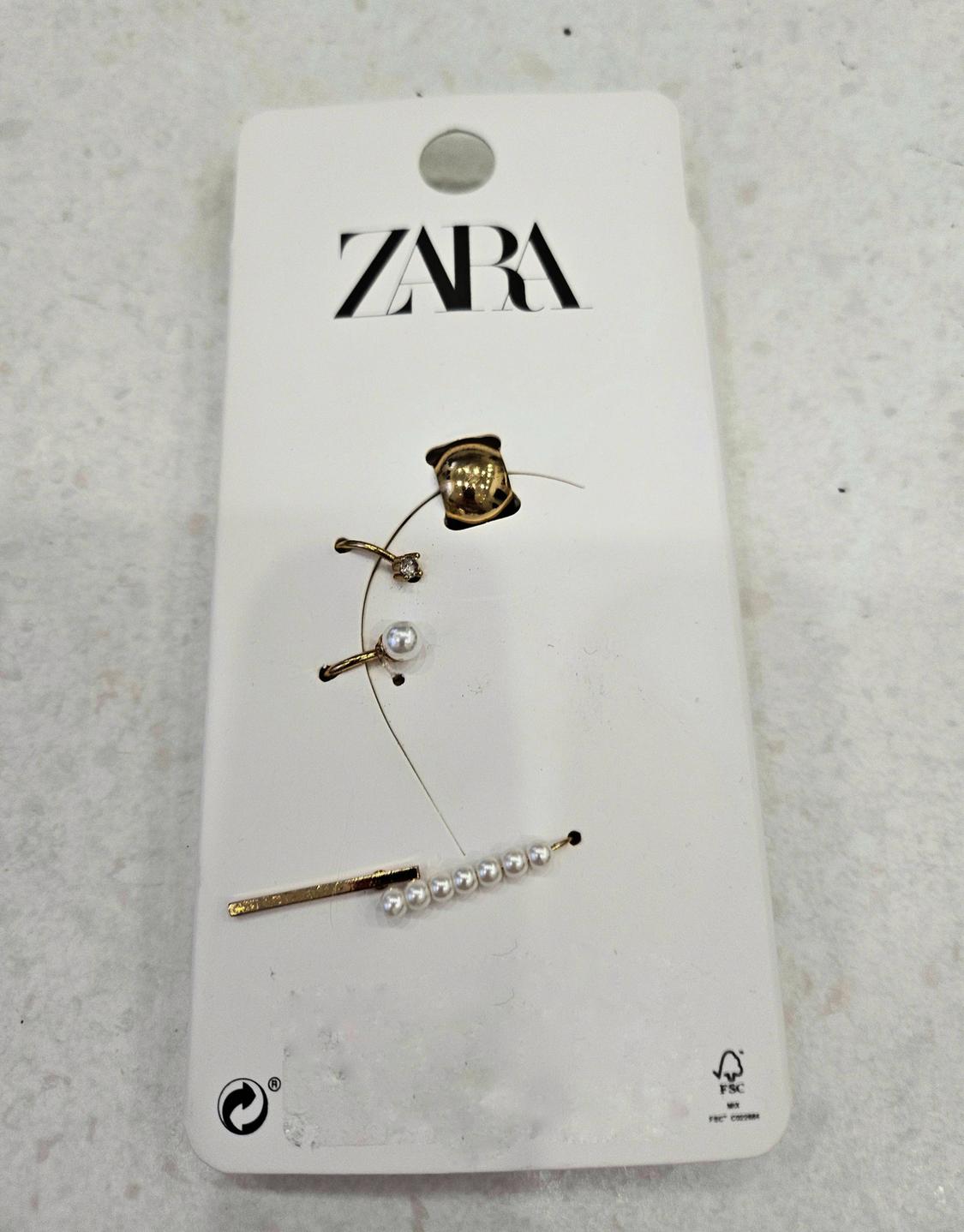 Λευκή κάρτα με το λογότυπο "ZARA" σε μαύρο, που δείχνει χρυσά σκουλαρίκια με πέρλες και λαμπερές λεπτομέρειες πάνω σε μια φωτεινή επιφάνεια.