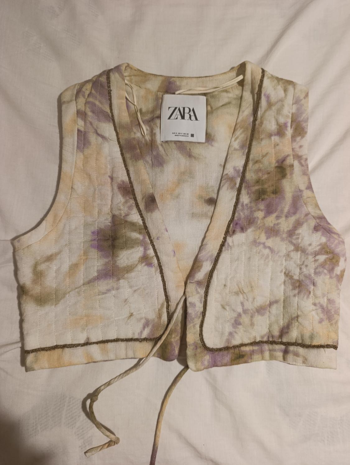 Cropped fabric gilet with a tie-dye print in beige and purple tones, visible "ZARA" label, on a white background.