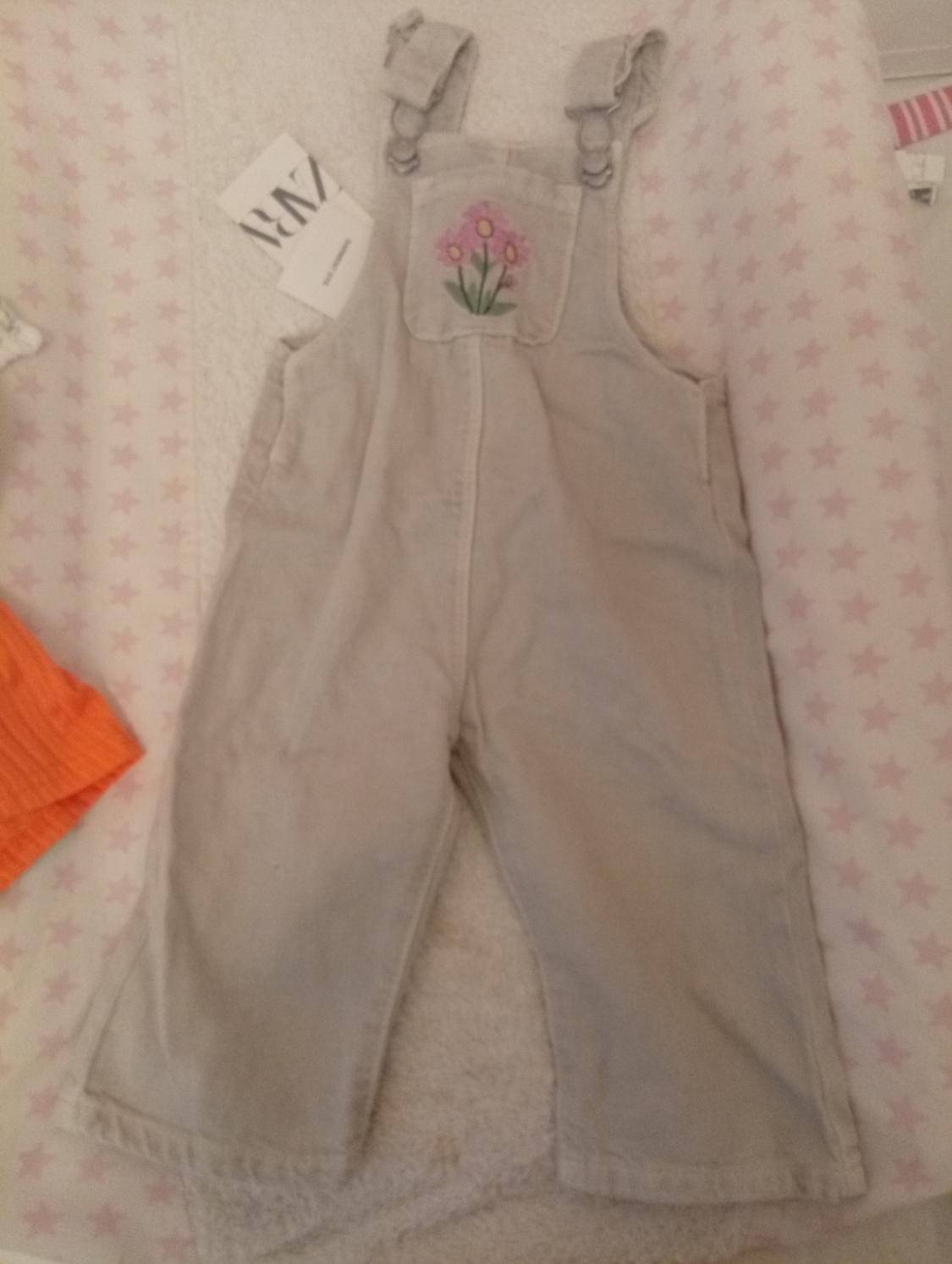 Beige baby dungarees with pink flower embroidery on the pocket. Background with pink star print and orange fabric on the left.
