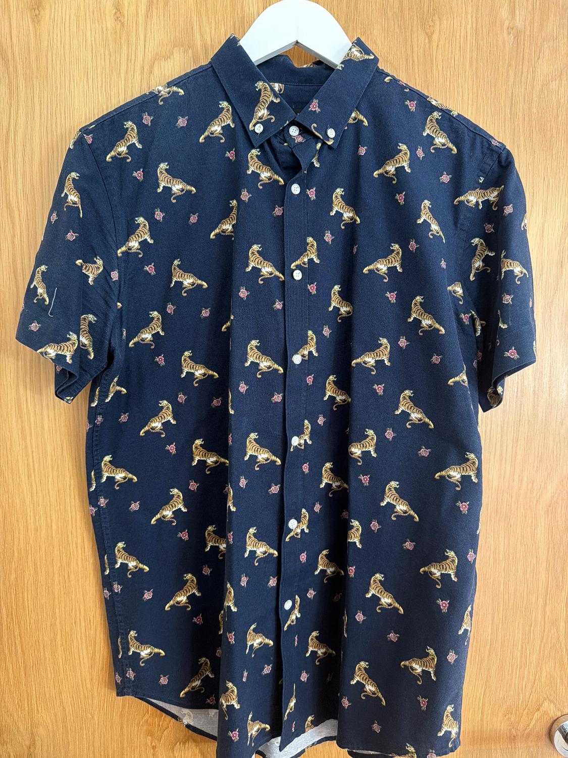 Navy short-sleeved shirt with a tiger and small floral print, hanging on a white hanger against a light wooden background.