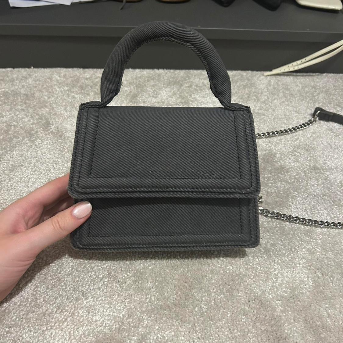 A black handbag with a metal chain is lying on a grey carpet. A hand is holding it. In the background, there are shelves with shoes.