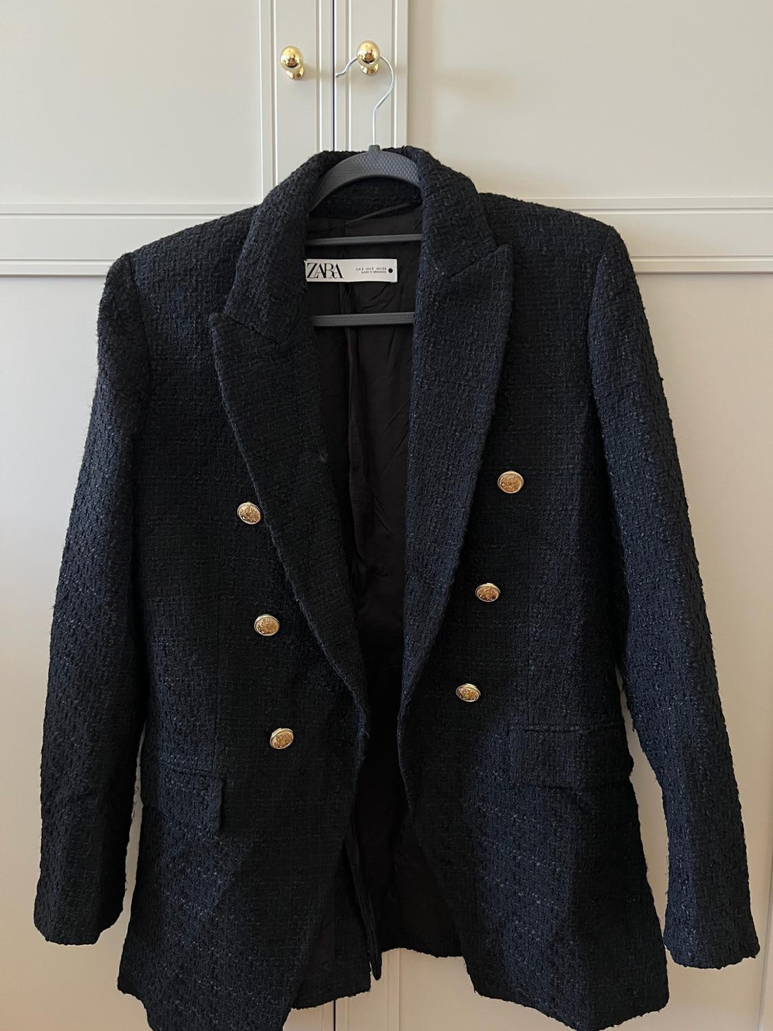 Textured black double-breasted blazer with gold buttons, hanging in a white wardrobe. Label with ZARA text. Relaxed style.