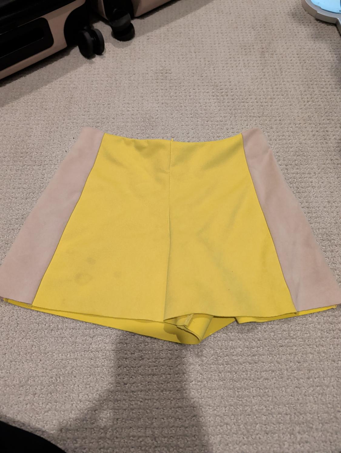 Yellow shorts with beige side panels on a beige rug, next to black and beige suitcases in the background.