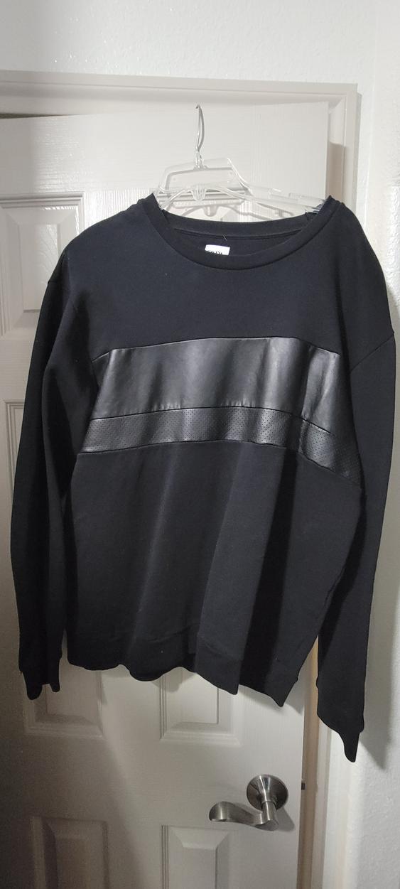 Black sweatshirt with a shiny material band on the chest, hanging on a white door with a clear hanger.
