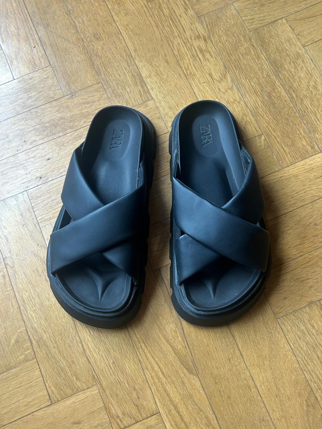 Black sandals with crossover straps and a chunky sole on a wooden floor. The text ZARA is visible on the insole.