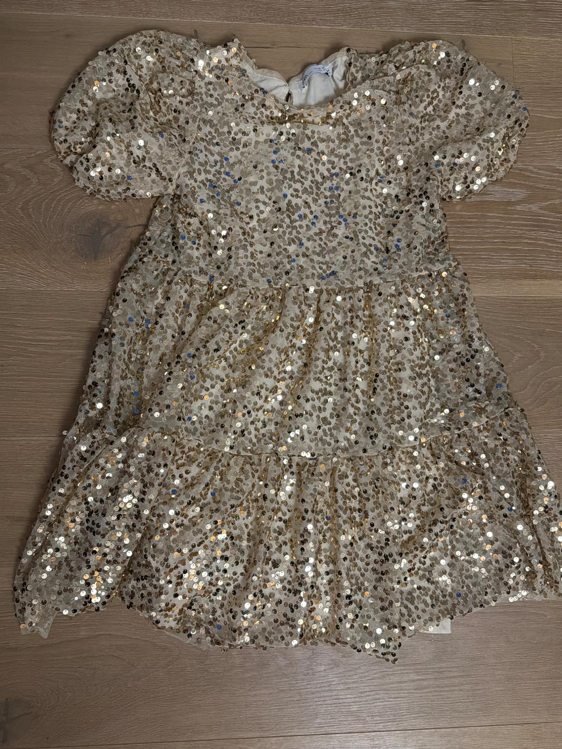 Child's dress with gold sequins on a beige background, with puffed sleeves, on a light wooden floor.