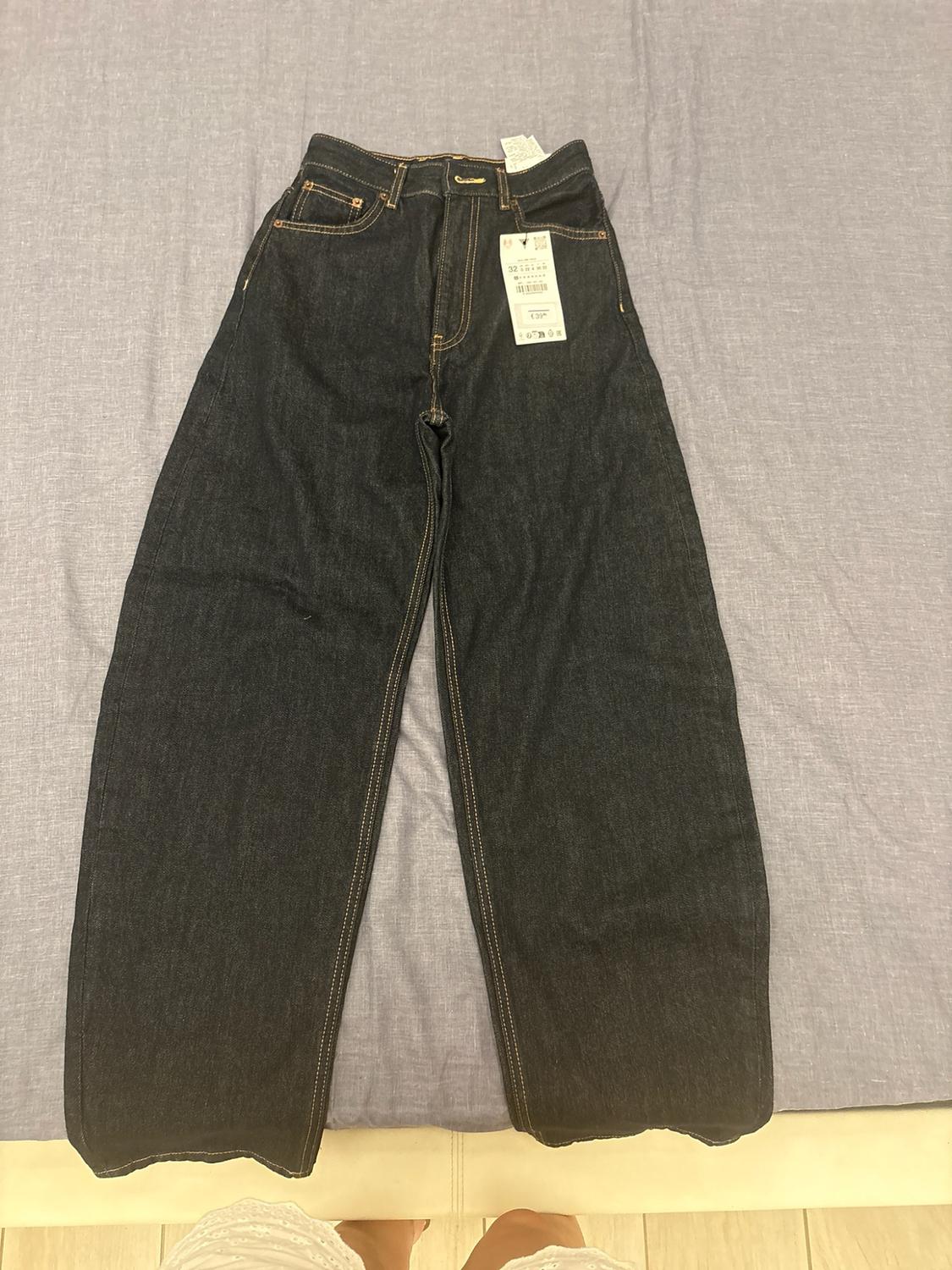 Wide dark-blue denim trousers on light-blue fabric. A label reads ‘32’ and ‘€39’. Bare feet are visible below.