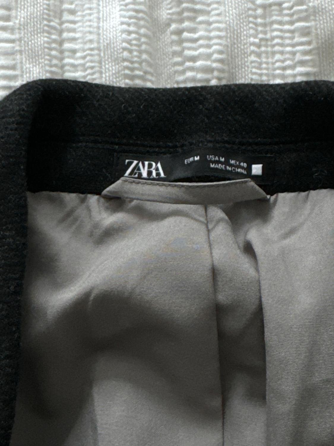 Black jacket label with grey interior, ZARA brand, size EUR M, USA M, MEX 40, made in China, on a woven white background.
