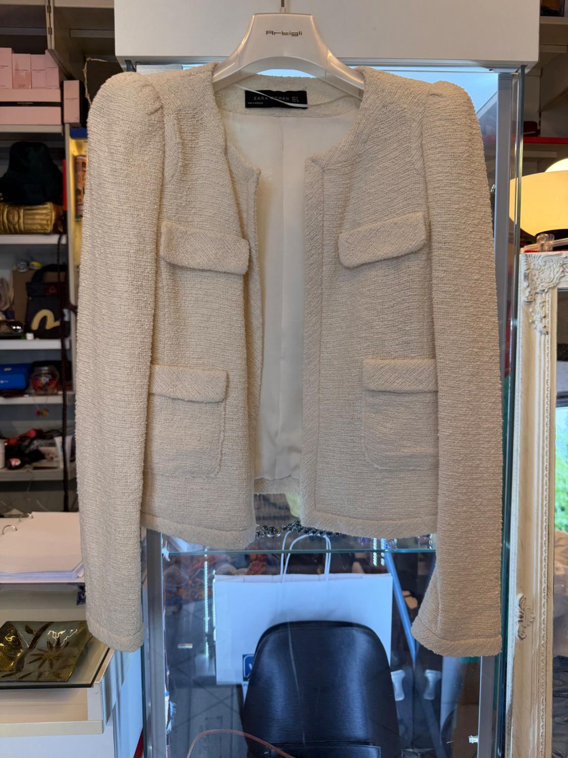 Cream-coloured textured jacket with four pockets, hanging on an Artigli hanger with a ZARA WOMAN label inside a shop.