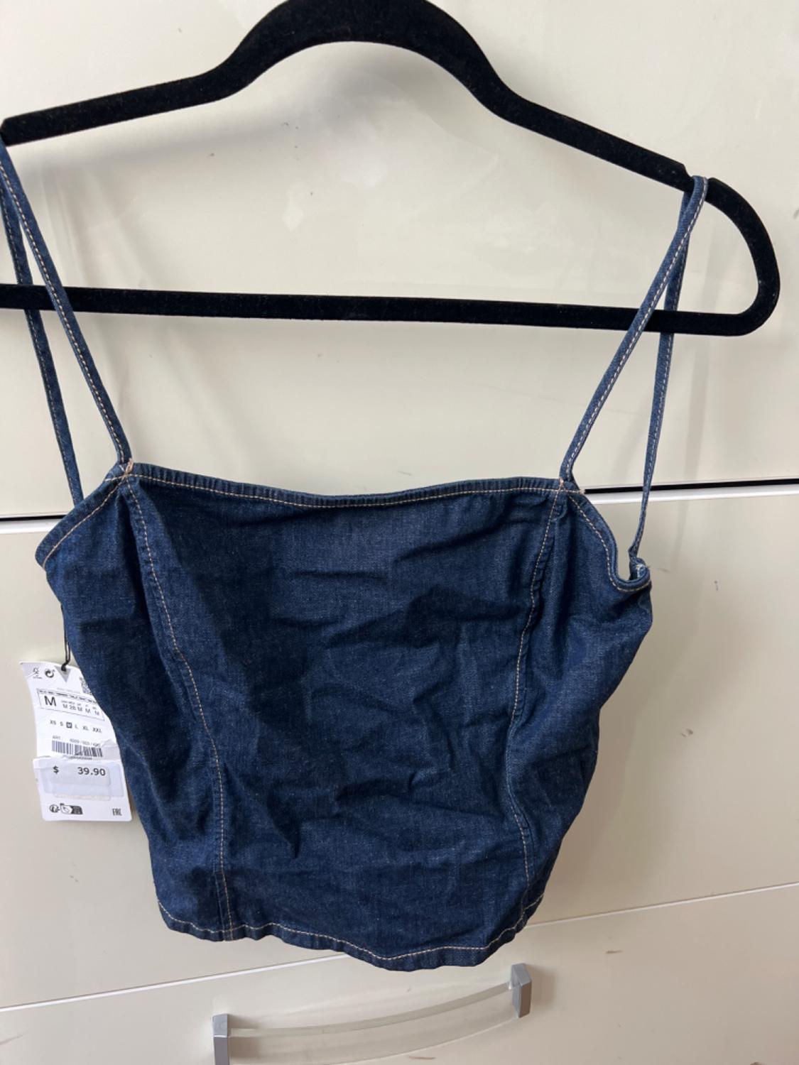 Blue denim spaghetti strap top hanging on a black hanger. Visible tag with price of $39.90 and size M. White background.