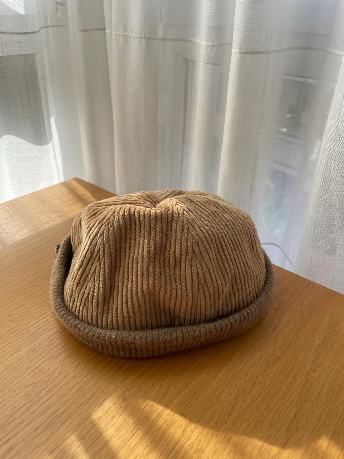 Beige corduroy beanie with a rolled brim on a wooden table, illuminated by natural light next to white curtains.