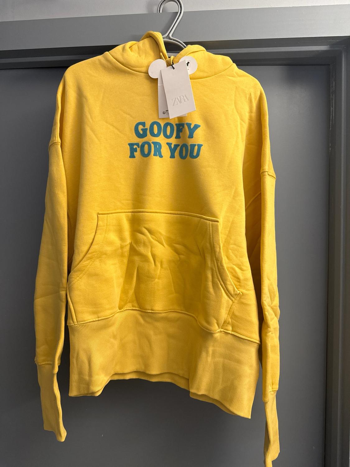 Yellow hoodie on a hanger against a grey background. It features a front pocket, a ZARA label, and the blue text GOOFY FOR YOU.