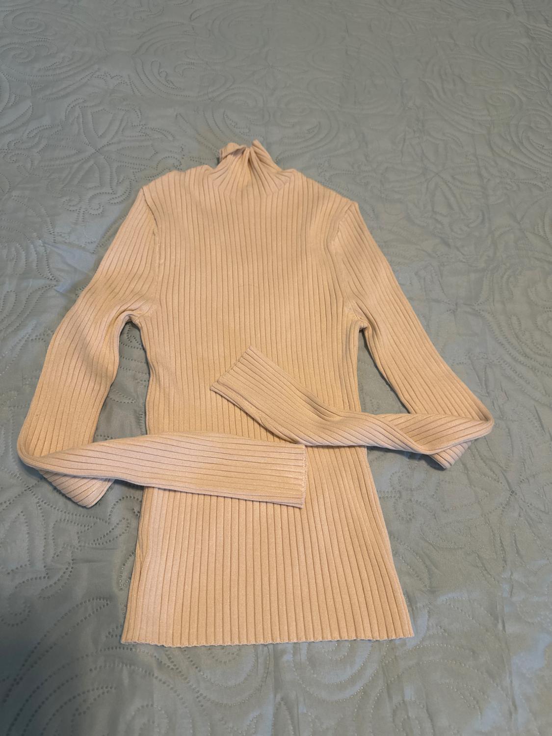 Light beige ribbed knit jumper with a high neck and long, crossover sleeves, laid out on a textured light blue bedspread.