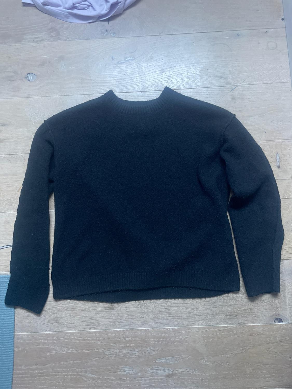 Black long-sleeved, round-neck jumper laid out on a light wooden floor.
