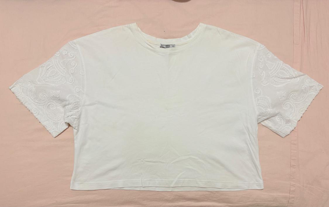 White short-sleeved T-shirt with embroidery on the sleeves, laid out on a light pink fabric background.