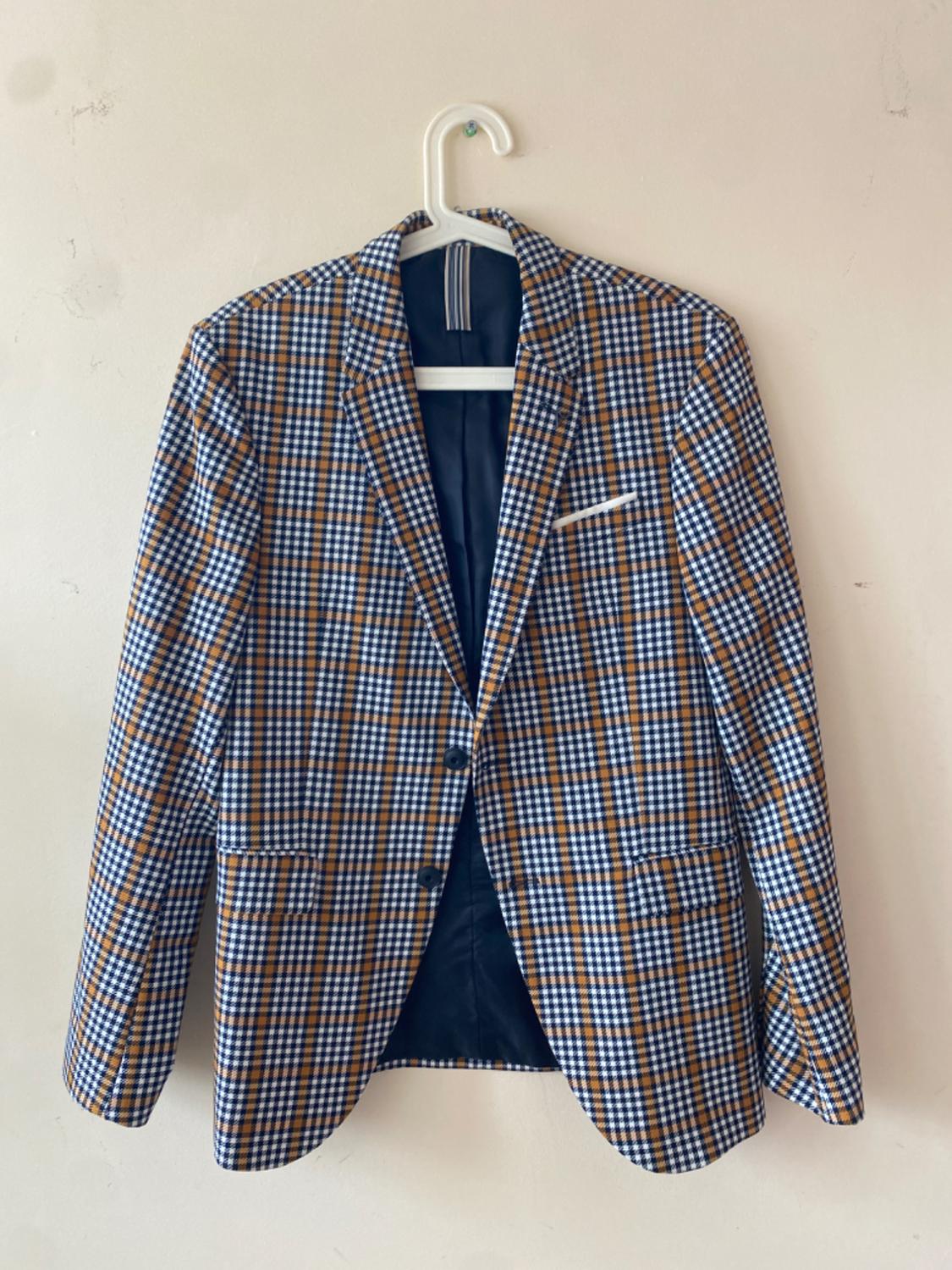 Brown and blue checked jacket hanging on a white hanger against a beige wall. Chest pocket visible.