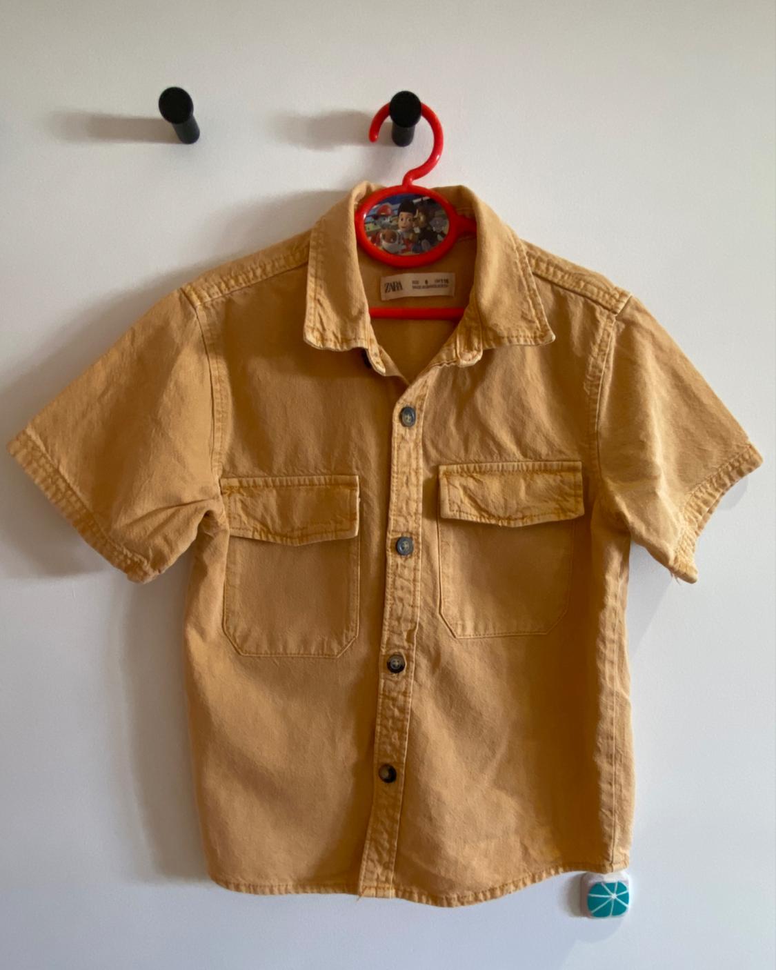 Beige short-sleeved shirt hanging on a red hanger with a children's design. It has two front pockets and dark buttons.