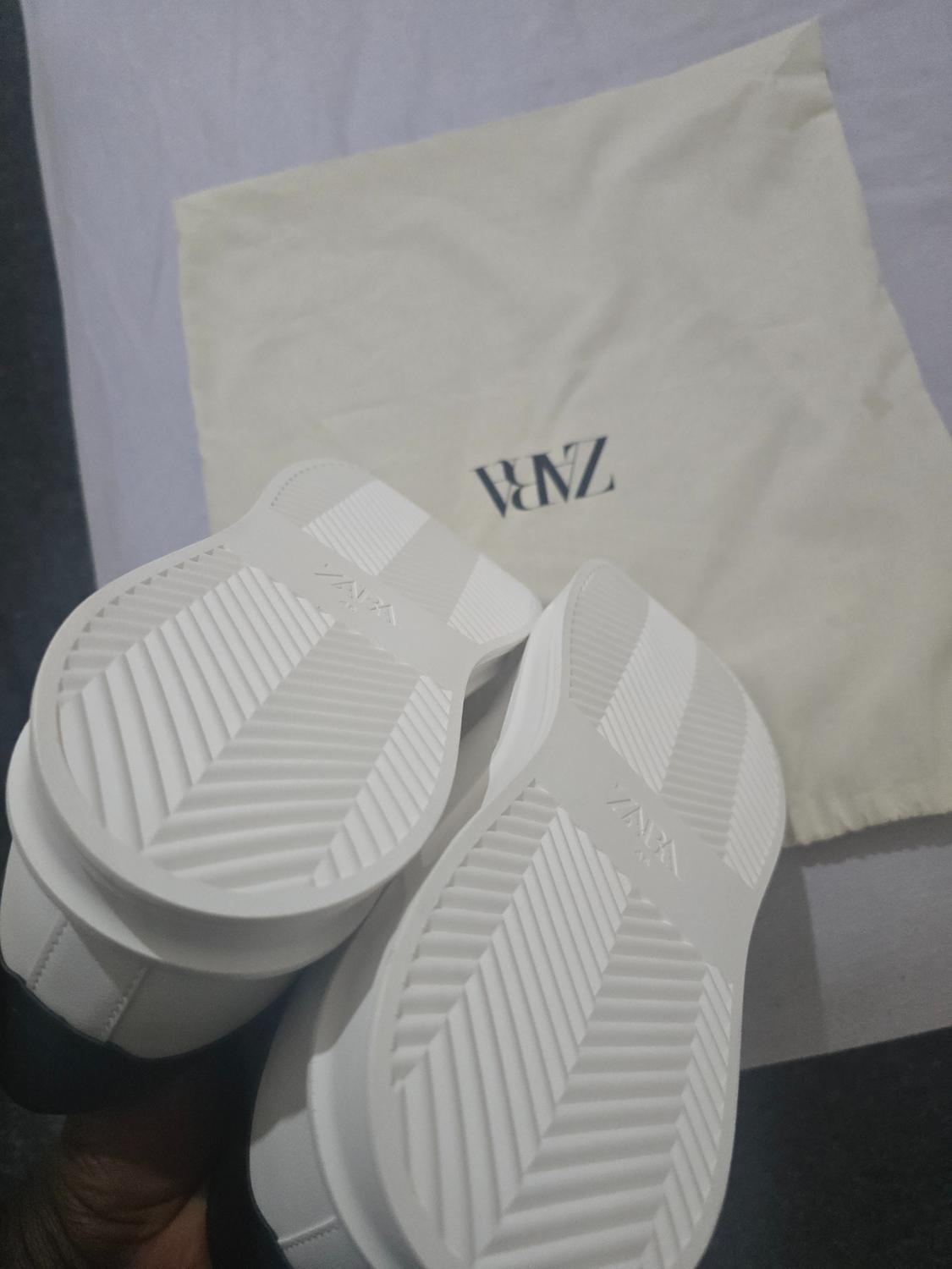 White trainers showing the soles with the text ZARA, held in front of a fabric bag bearing the word ZARA.