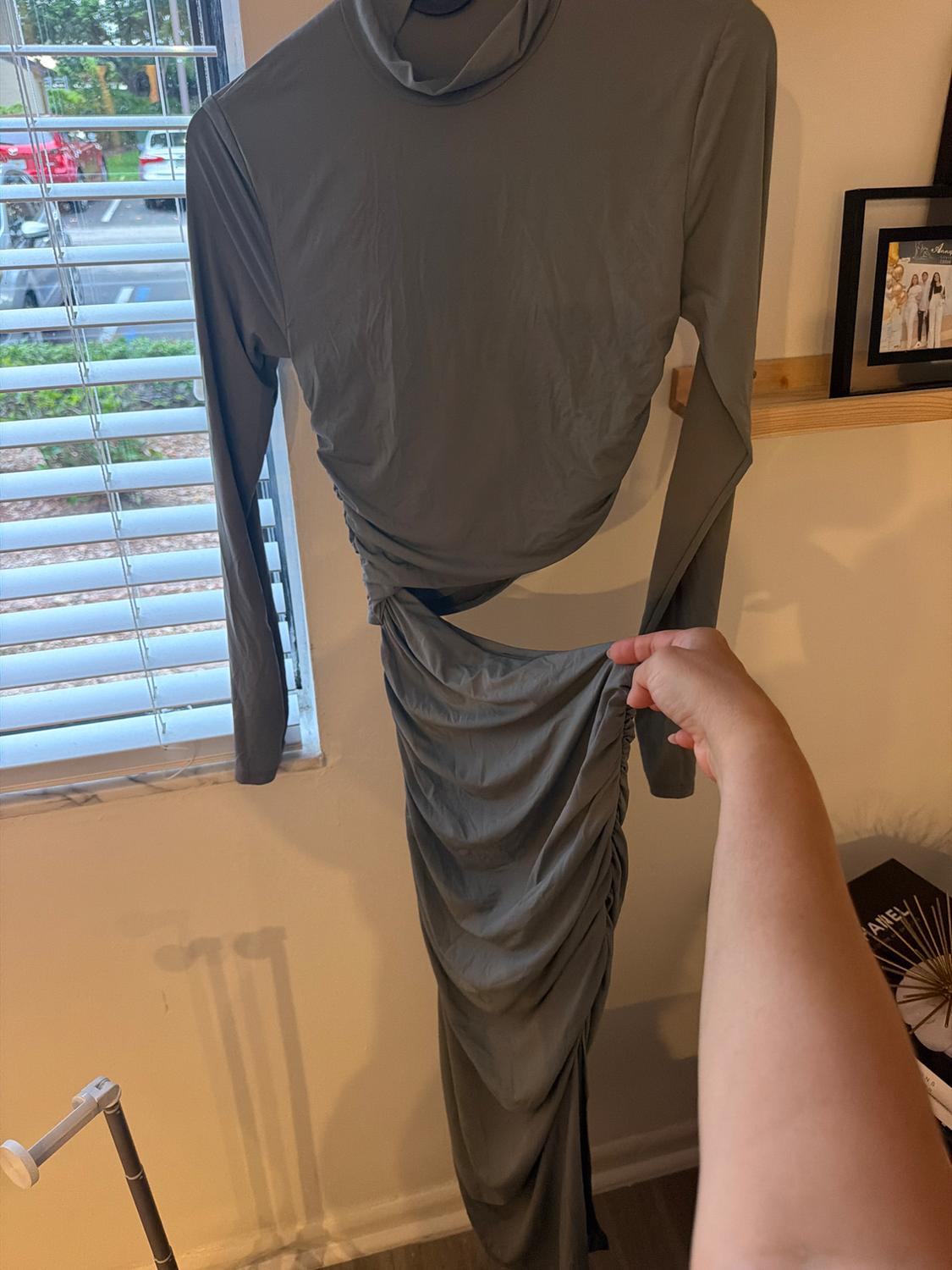Long-sleeve gray dress with a high neck, waist cutout, and gathered skirt, hanging next to a window. An arm holds the fabric.