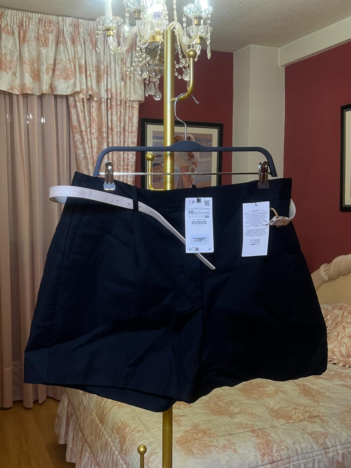 Black shorts hanging on a gold chandelier in a bedroom with patterned curtains and bedspread. Label visible.