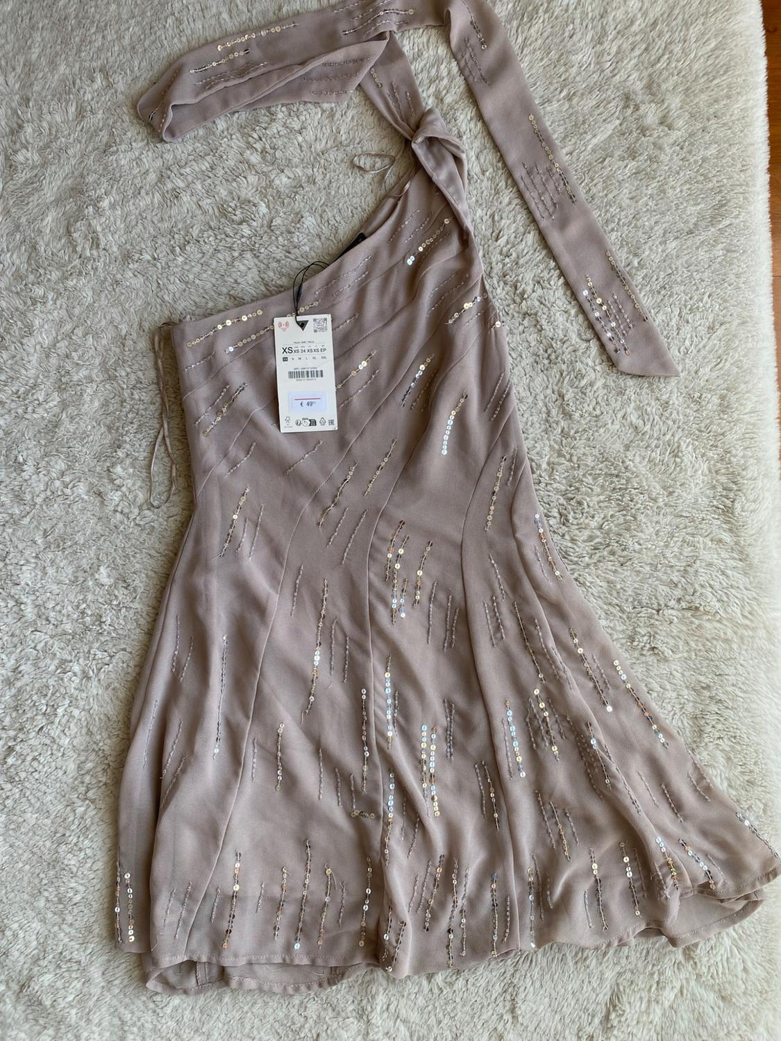 Beige asymmetrical dress with silver sequins on a plush rug. Label with text: XS XS 24 XS XS EP, €49.95.