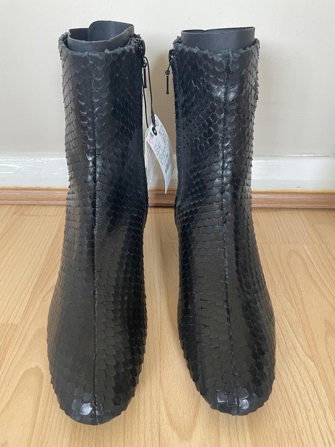 Black boots with a scale texture, front view on a light wooden floor. Tags hanging from the side zips.