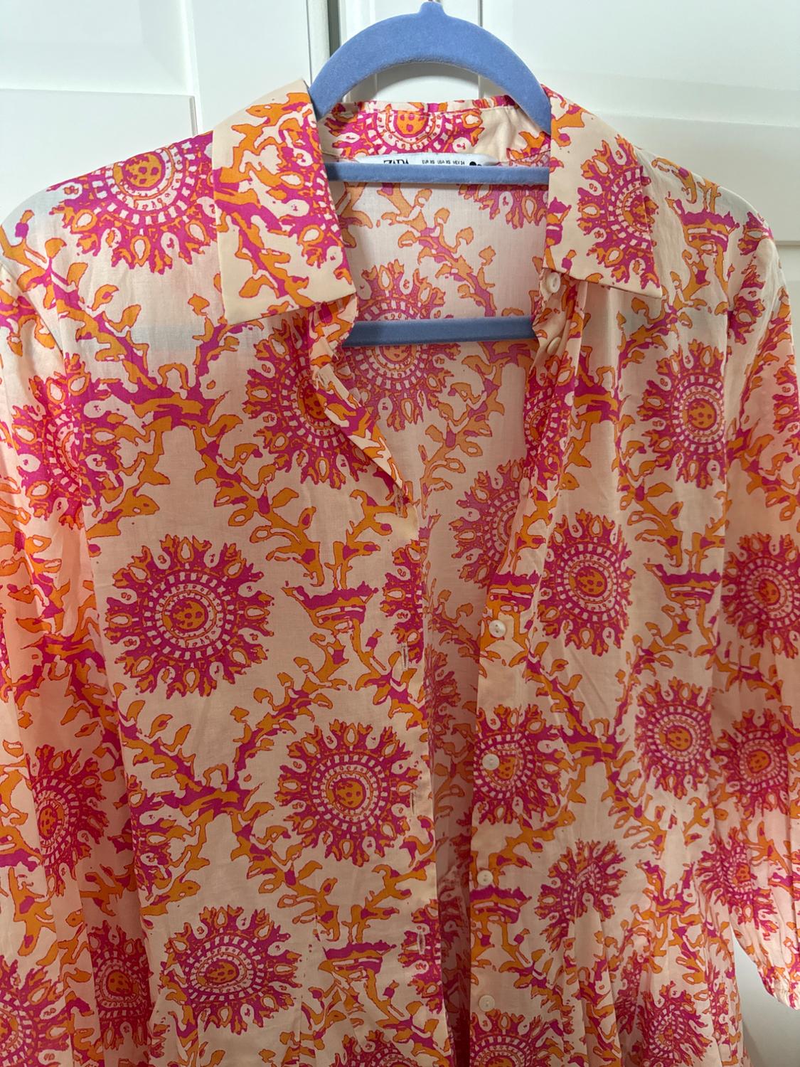 Pink and orange patterned shirt hanging on a blue hanger in front of white doors. Text: ZARA EUR XS USA XS MEX 24.