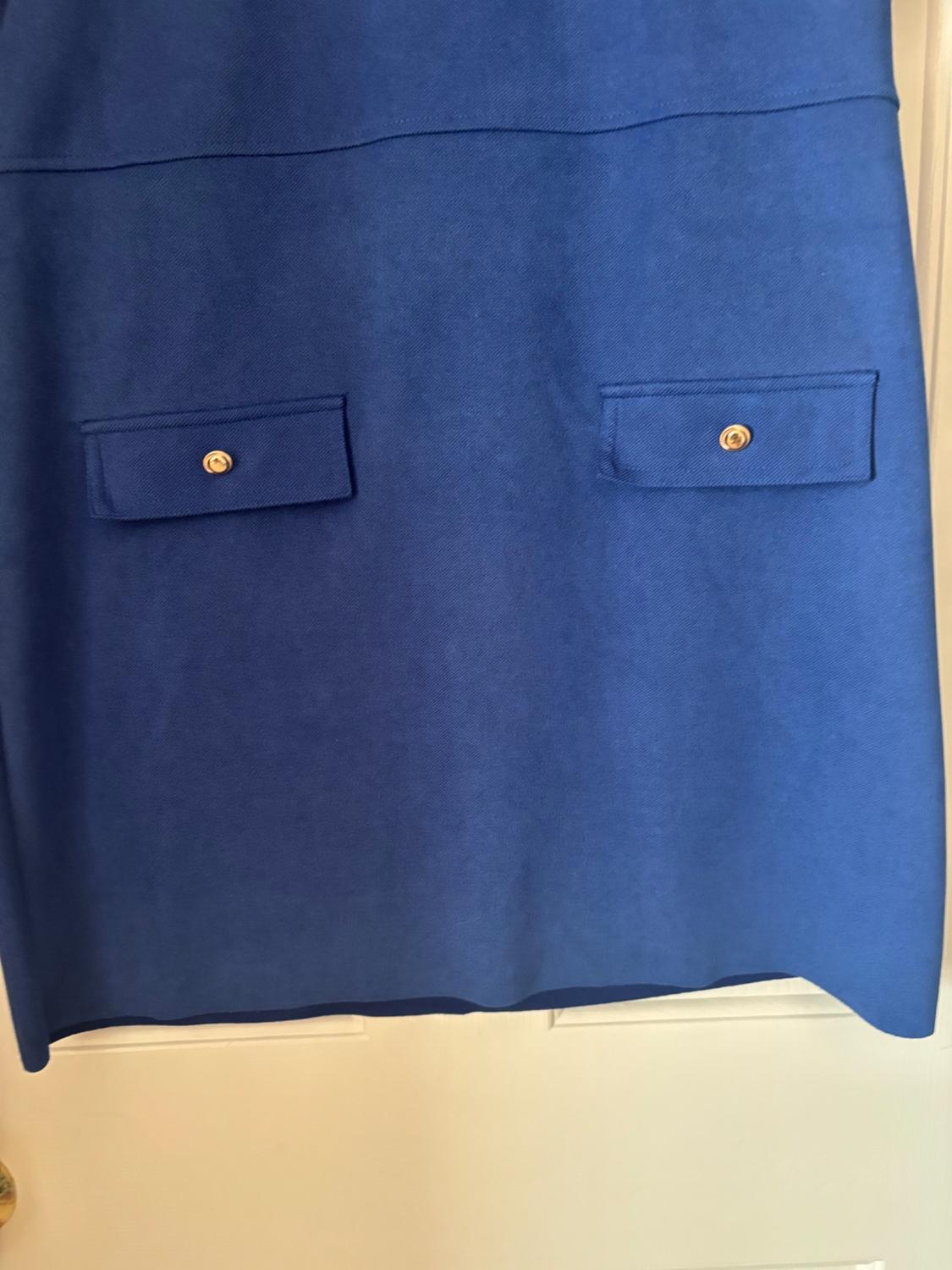 Blue skirt with two front flap pockets and gold button, hanging on a white door.