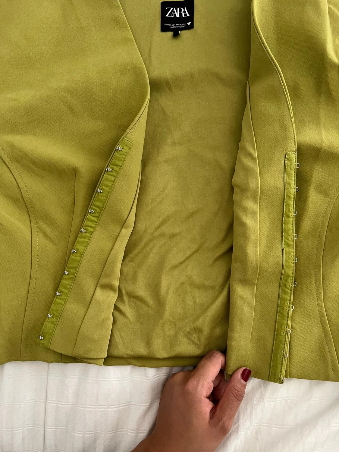 Close-up of a green garment with hook-and-eye closures on a white fabric. A hand with red-painted nails is holding it. The text reads: ZARA.