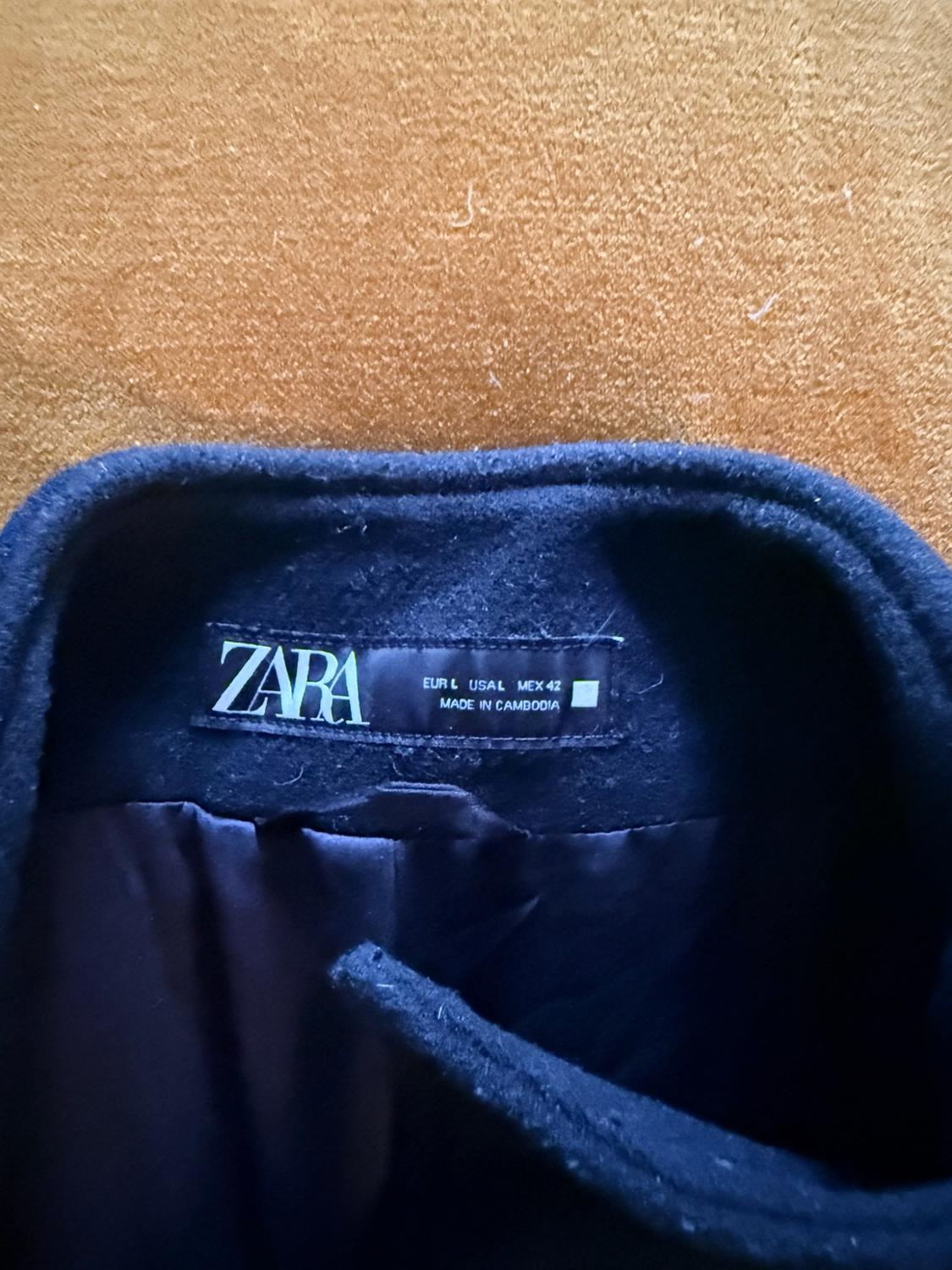 Close-up of a label on the collar of a dark garment against a brown background. Text: ZARA EUR L USA L MEX 42 MADE IN CAMBODIA.