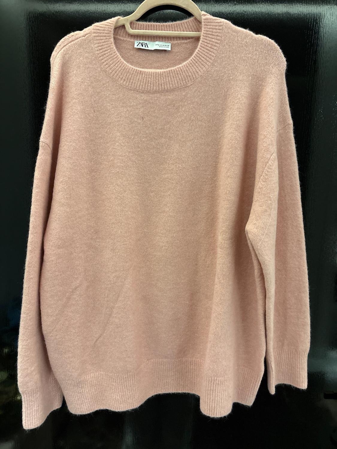 Light pink knitted jumper hanging on a wooden hanger, with a round neck and a visible Zara label, black background.